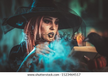 Witch with awfully face in creepy surroundings and smoky green background reading recipe of magic drink.