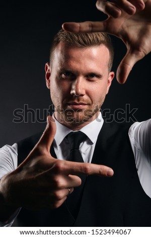 young handsome man in white shirt, black vest making frame with his hands on black background, close up photo. isolated black background
