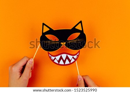 Funny face of monster with cat mask and vampire smile on orange background. Female hands are holding paper photo props on canvas. Party carnival accessories for celebration happy halloween.