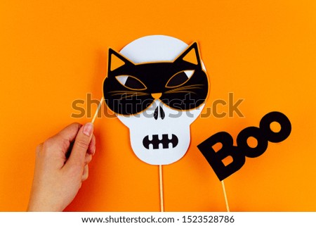 Funny face of skull monster with cat mask and black inscription boo on orange background. Female hand is holding paper photo props on canvas. Party accessories for celebrating happy halloween.