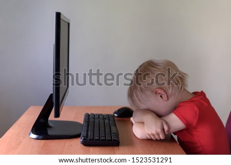 Tired little blond boy sleeping on a table under the monitor PC. Internet and preschooler