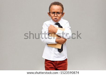 Studio picture of greedy nerdy dark skinned elementary pupil wearing school uniform and eyeglasses holding tight copybook and exclaiming. Childhood, education, learning and lifestyle concept