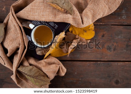 Cropped photo of a white cup with tea and a violet sweater strewn with orange maple leaves on a wooden table. The concept of comfort, coziness and hygiene. 