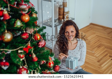 Indoor portrait of a beautiful brunette woman at Christmas time, sitting on the floor near Christmas tree and holding gift box.