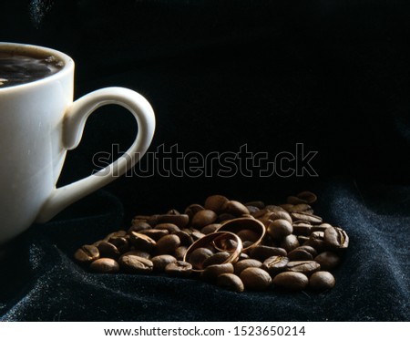 White cup of coffee on a black background and coffee beans. Two gold rings lie in the grains. Concept - morning breakfast for honeymooners