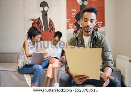 Young African American man thoughtfully opening envelope with colleagues working on background in modern co-working space