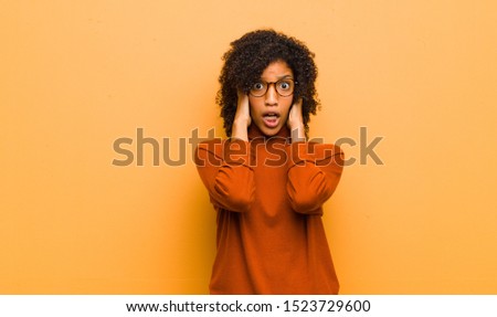 young pretty black woman looking unpleasantly shocked, scared or worried, mouth wide open and covering both ears with hands against orange wall