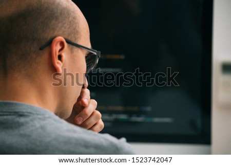 Bald caucasian programmer in glasses is sitting behind the desk, in front of black monitor, looking closely, analysing code lines, thinking through. He's very attentive and focused. Close up.