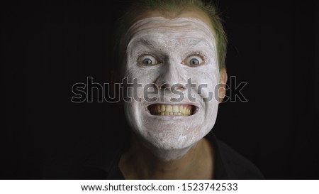 Clown Halloween man portrait. Close-up of an crazy, evil clowns face. White face makeup. Green hair. Scary laugh. Attractive model in Halloween costume. Dark background