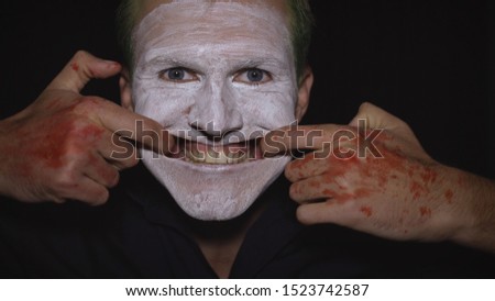 Clown Halloween man portrait. Close-up of an crazy, evil clowns face. White face makeup. Green hair. Scary smile with fingers. Attractive model in Halloween costume. Dark background