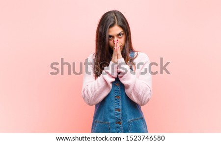 young  pretty woman feeling worried, hopeful and religious, praying faithfully with palms pressed, begging forgiveness against pink background.