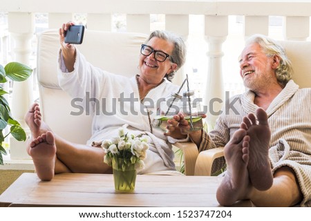 couple of seniors or mature people in a resort spa in their hotel or house clinking with their cocktail and taking a salfie with her phone - having fun with feet on the table looking between they