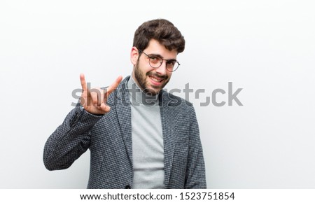 young manager man feeling happy, fun, confident, positive and rebellious, making rock or heavy metal sign with hand against white wall