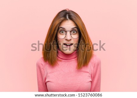 young pretty woman feeling terrified and shocked, with mouth wide open in surprise against pink background