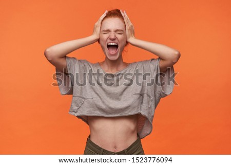 Portrait of stressed young readhead female in grey t-shirt and green shorts isolated over orange background, holding head with hands and screaming loud with wide mouth opened