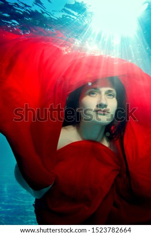 Underwater portrait of an extraordinary girl. She poses underwater with a red cloth over her head, against the sun's rays from the surface. Close up. Surrealism. Underwater photography