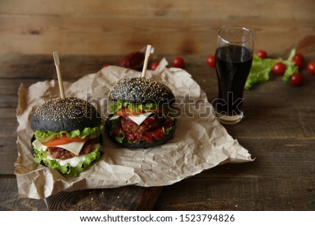 Two black sandwich, hamburger or burger with beef or chicken, tomatoes, onions, cheese, lettuce and a sesame bun with drink of cola in a glass on a paper on a wooden table. Fast food. Copy space.