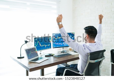 Freelance stock broker celebrating successful trading while sitting with arms raised and working at home Royalty-Free Stock Photo #1523848127