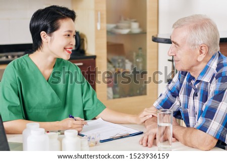 Smiling Vietnamese nurse touching hand of senior patient and filling his medical card when visiting him at home