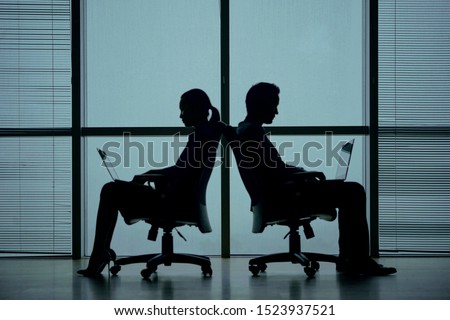 Silhouettes of two business people sitting back to back in office chairs against big panoramic window and working on laptop