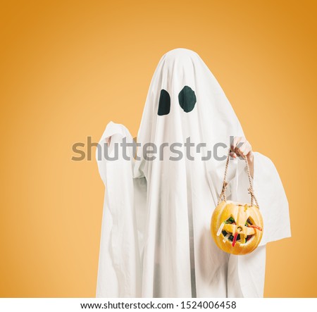 Cute white ghost with black eyes holding pumpkin basket with sweet candy on orange background.