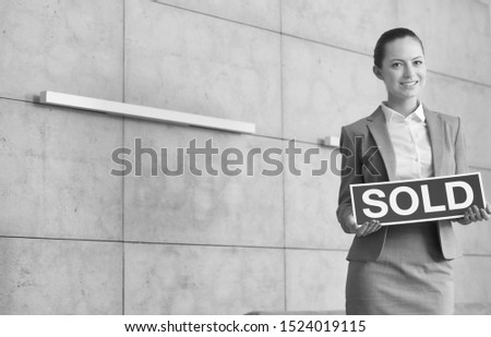 Black and white photo of confident young saleswoman holding sold placard while standing against wall in apartment