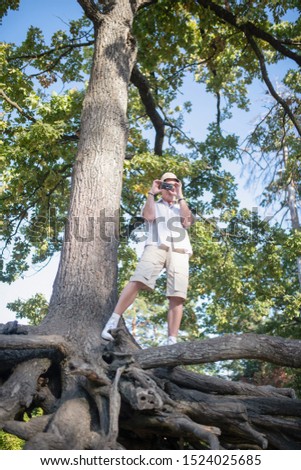 On big tree. Mature man wearing shorts standing on big tree and taking photo of forest