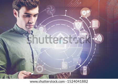 Serious young man in green shirt with laptop using futuristic smart home interface in blurry kitchen. Concept of automation and IoT. Toned image double exposure