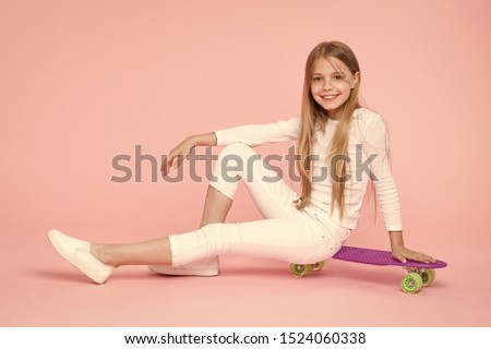 Riding board in style for years to come. Happy little child sitting on board deck on pink background. Cute small skater smiling with violet penny board. Taking pleasure in skateboarding on her board.