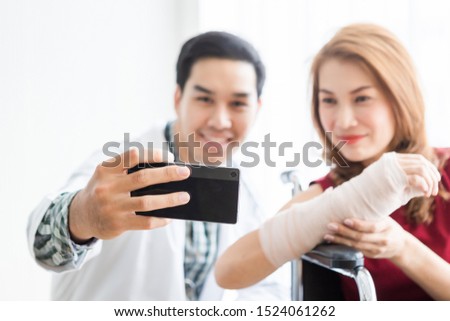 Selfie of smiling man doctor with a female patient wear arm splint for better healing sit in a wheelchair Abstract blur with focus using a smartphone In the room hospital background.