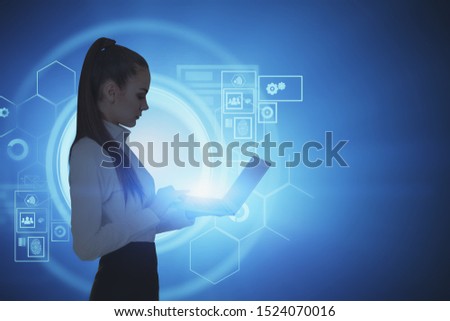 Side view of serious young businesswoman standing with laptop over blue background with futuristic HUD interface. Concept of coding and software. Toned image mock up