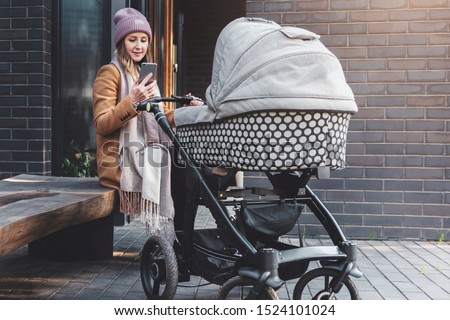 Woman with baby stroller walking in local area. Young mother with sleeping child in carriage sitting on bench in recreation park zone using smartphone for online communication. Girl texting messages.