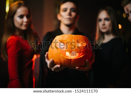 Group of friends celebrating Halloween. Guy wearing zombie costume holds pumpkin on his outstretched arm. Two beautiful girls looking at pumpkin. People's faces are blurred, camera focuses on pumpkin