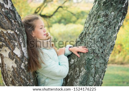 Pensive girl in blue down jacket leaning on birch tree on an autumn day