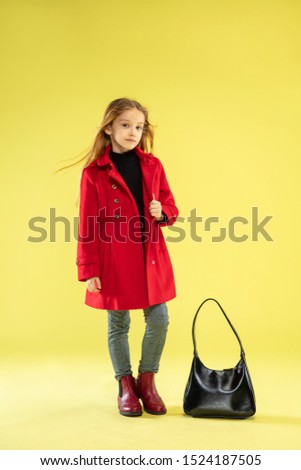 A full length portrait of a bright fashionable girl in a red raincoat with black bag posing on yellow studio background. Looks happy. Autumn and spring fashion for kids. Cute stylish blonde girl.