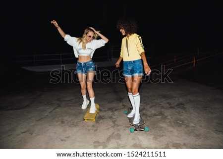 Image of two stylish girls in streetwear smiling and riding skateboards at night outdoors