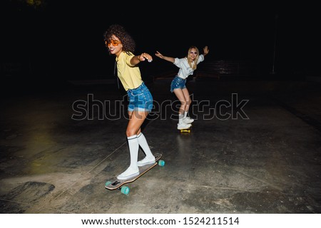Image of two multinational girls in streetwear smiling and riding skateboards at night outdoors