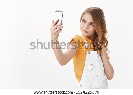 Sassy glamour cute pretty little girl with blond hair hold smartphone, taking selfie posing feminine and silly, pouting stare confident, mimicking adult women, stand white background