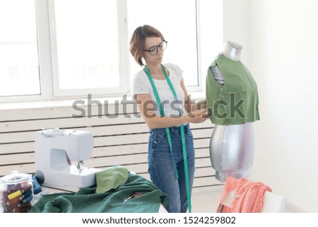 Young pretty girl seamstress designer with glasses and a measuring tape makes a new product with the help of a green cloth and a tailor's dummy. Concept of sewing workshop.