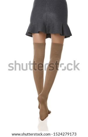 Medium back bottom shot of a female figure in the gray bell-shaped skirt and beige shadow-proof stockings with closely rim on the top. The girl on tiptoe is posing on the white background. 