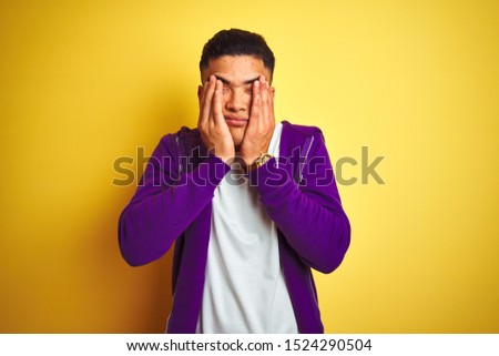 Young brazilian man wearing purple sweatshirt standing over isolated yellow background rubbing eyes for fatigue and headache, sleepy and tired expression. Vision problem