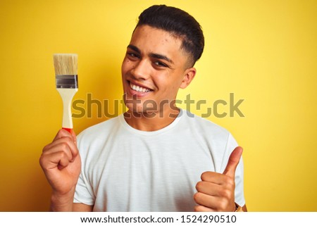 Young brazilian painter man holding brush standing over isolated yellow background happy with big smile doing ok sign, thumb up with fingers, excellent sign