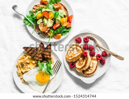 Savory and sweet breakfast - avocado, cherry tomatoes grilled  bread salad, fried egg with hummus and rye croutons and pancakes with greek yogurt and raspberries on light background, top view  