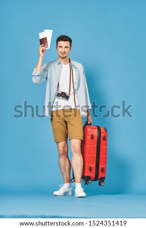 A man in shorts with a passport with airplane tickets in his hand a red suitcase and a blue background