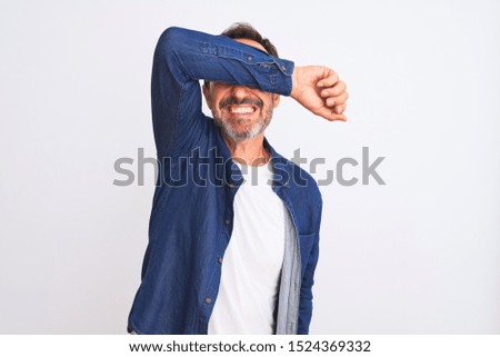 Middle age handsome man wearing blue denim shirt standing over isolated white background covering eyes with arm smiling cheerful and funny. Blind concept.