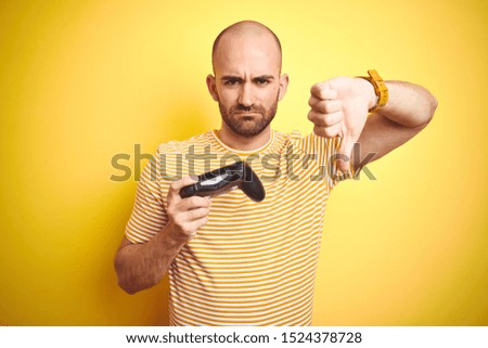 Young man playing video games using joystick gamepad over isolated yellow background with angry face, negative sign showing dislike with thumbs down, rejection concept
