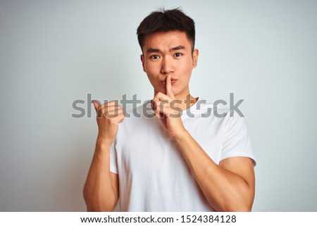 Young asian chinese man wearing t-shirt standing over isolated white background asking to be quiet with finger on lips pointing with hand to the side. Silence and secret concept.