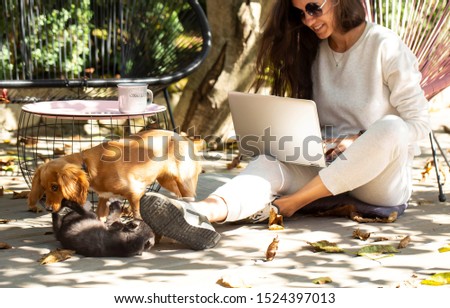 Portrait Of Beautiful Smiling Woman Typing On Notebook Keyboard While Working Online Using Laptop Computer. Female Sitting On Terrace At Cafe With Her Pets And Pc On Her Knees. Business People