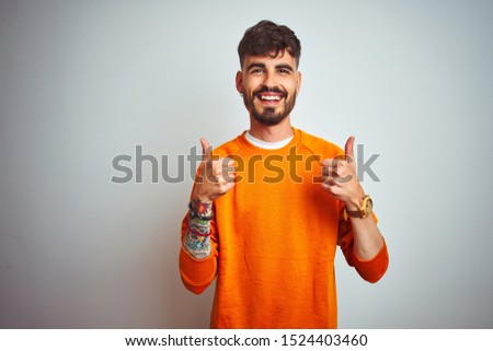Young man with tattoo wearing orange sweater standing over isolated white background success sign doing positive gesture with hand, thumbs up smiling and happy. Cheerful expression and winner gesture.