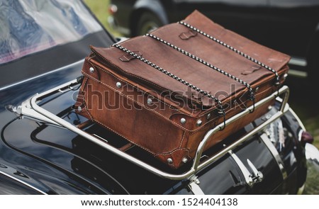 Close up of a vintage brown travel suitcase, strapped on a shiny chrome frame on the back of a black vintage car.  